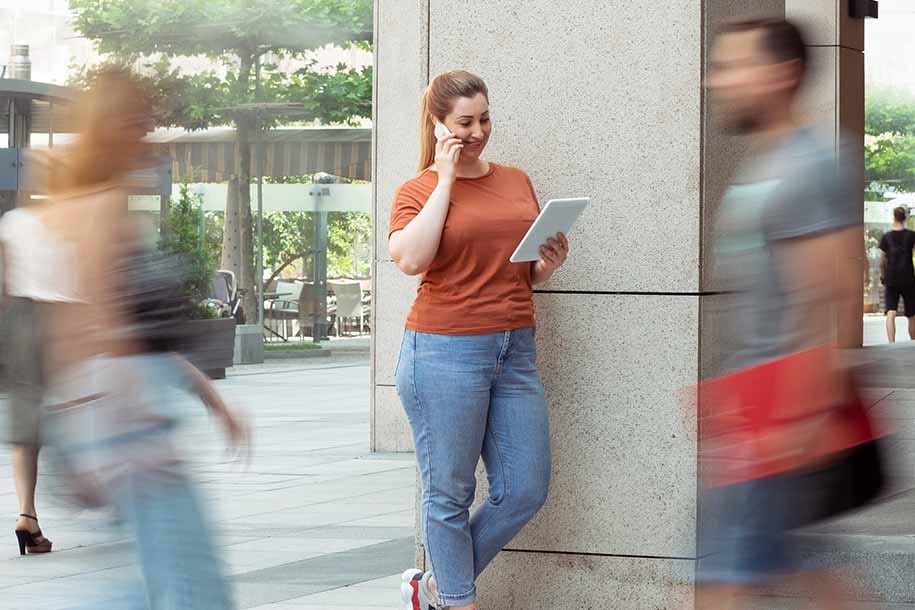 Vrouw in oranje shirt en spijkerbroek tegen betonnen pilaar, tablet in hand, omringd door bewegende voetgangers in stedelijke omgeving