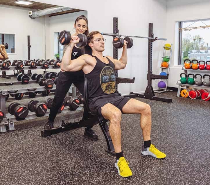 Personal trainer ondersteunt man bij schouderoefening met halters in fitnessruimte