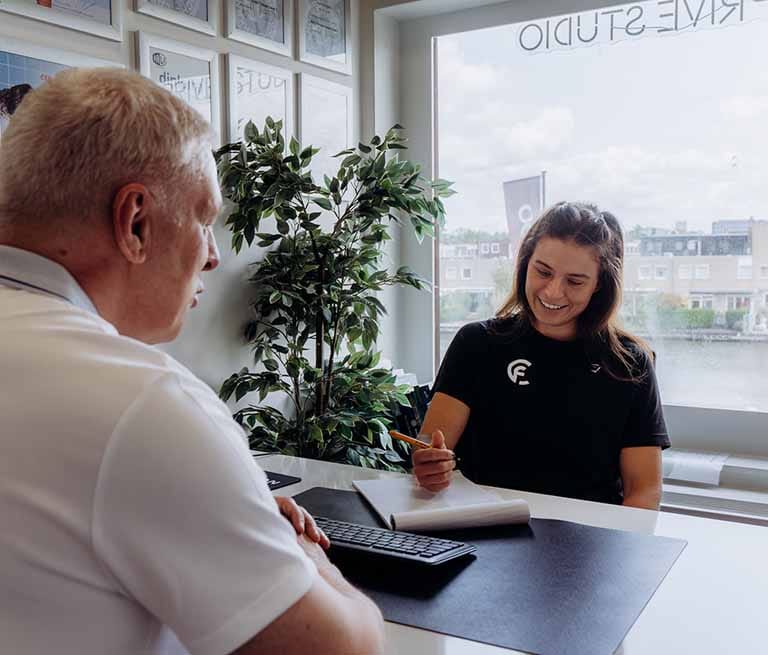 Trainer voert intakegesprek met cliënt aan bureau in modern kantoor met uitzicht op water