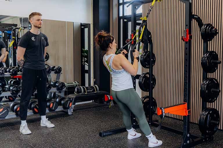 Personal trainer begeleidt vrouw bij kabeltraining in fitnessruimte met halterrek