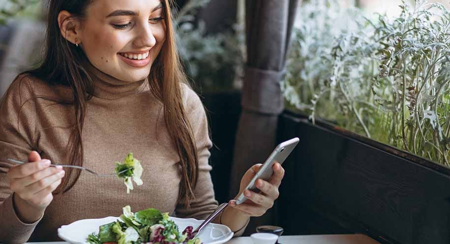 Vrouw eet gezonde salade terwijl zij op haar telefoon kijkt in een groene omgeving