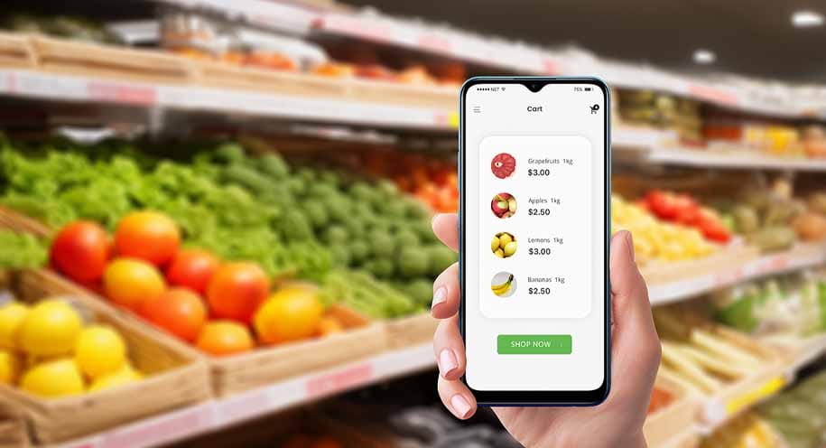 Hand houdt smartphone met winkelwagen app vast in supermarkt met groente- en fruitafdeling op achtergrond