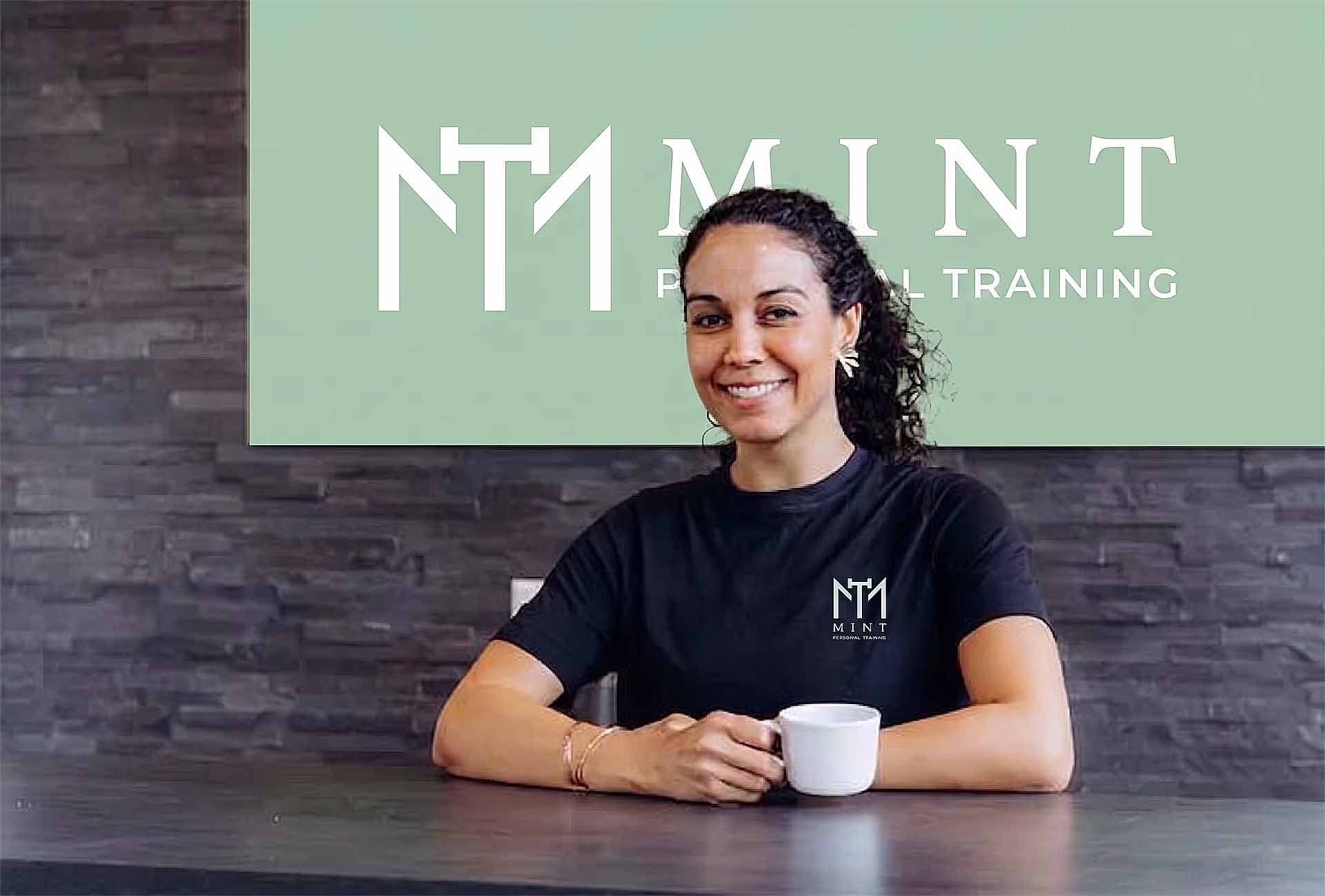 Vrouw in zwart Mint-shirt zit aan tafel met kopje koffie, lachend naar camera, achter haar groen bord met logo