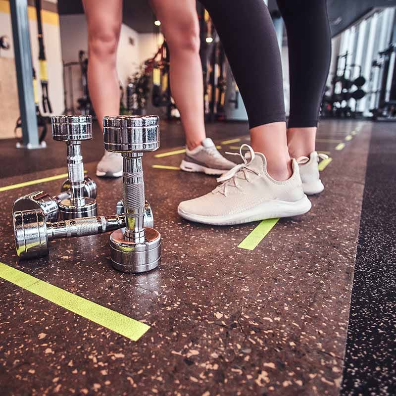 Twee personen in sportkleding en witte sneakers staan naast zilveren dumbbells op een sportmat met gele lijn in een fitnessruimte