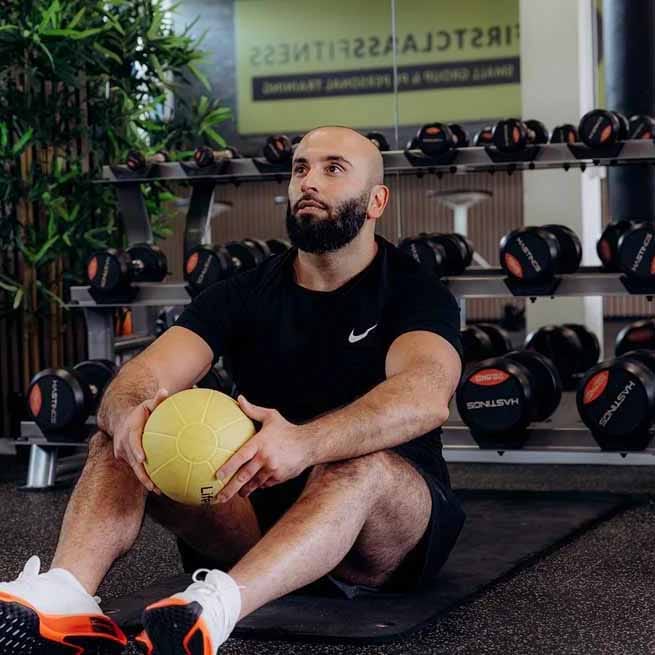 Man in zwart Nike-shirt zit op fitnesmat met gele medicine ball in personal training studio met halters op achtergrond