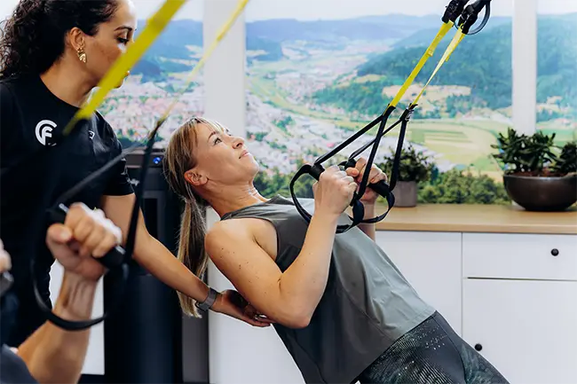Vrouw doet training aan TRX-banden onder begeleiding van een trainster in een studio met berglandschap op de achtergrond