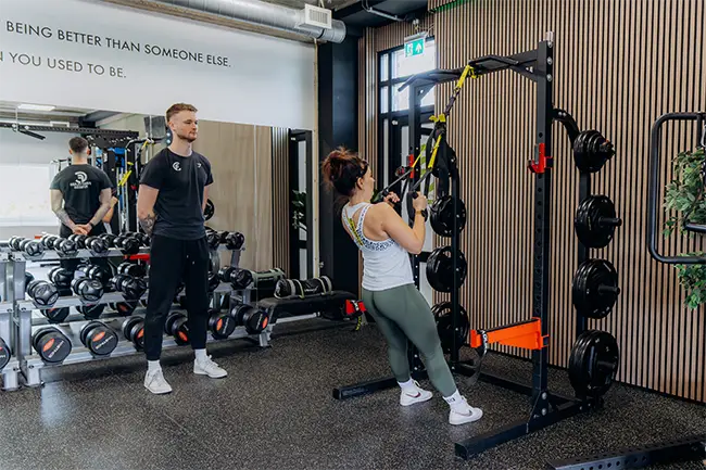 Vrouw traint onder begeleiding van personal trainer in moderne fitnessruimte met gewichten en trainingsapparatuur