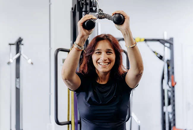 Vrouw met roodbruin haar houdt twee zwarte halters boven haar hoofd en lacht in een moderne trainingsstudio