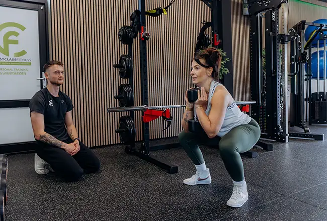 Personal trainer begeleidt vrouw bij squat-oefening in trainingsstudio met apparatuur op achtergrond