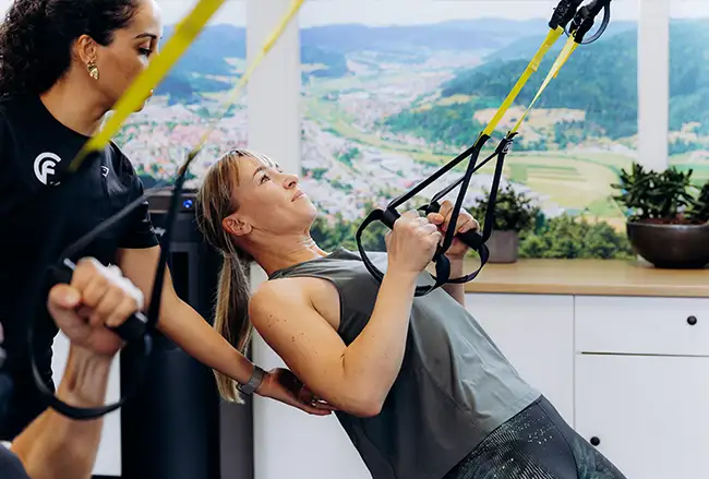 Vrouw doet suspension training oefening onder begeleiding van trainer in studio met uitzicht op berglandschap