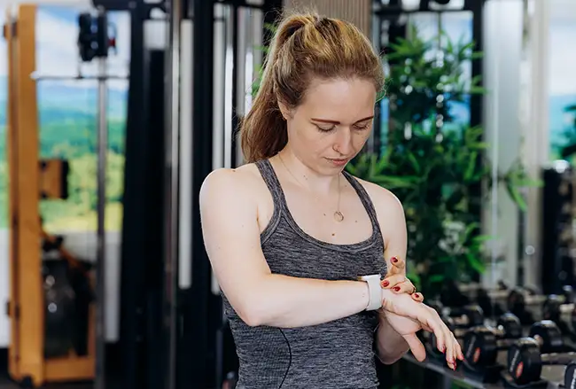 Vrouw in sportkleding controleert hartslagmeter aan haar pols in een trainingstudio met apparatuur en groen