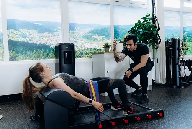 Personal trainer begeleidt vrouw bij training op apparaat met berglandschap op achtergrond